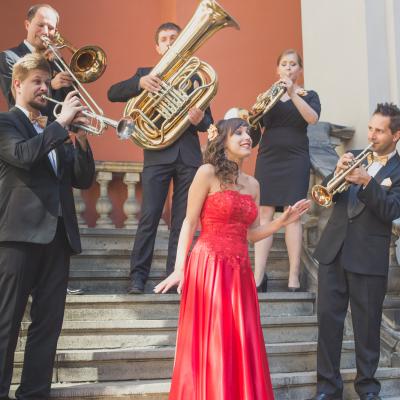 Członkowie zespołu Masterbrass, ubrani elegancko ze złotymi dodatkami wraz z solistką ubraną w długą, czerwoną suknię, ze złotym kwiatkiem wpiętym we włosy, grający na schodach dziedzińca Ogólnokształcącej Szkoły Baletowej im. Olgi Sławskiej Lipczyńskiej.