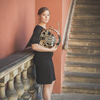 Waltornistka, ubrana w czarną sukienkę, stojąca bokiem ze swoim instrumentem, na tle schodów dziedzińca Ogólnokształcącej Szkoły Baletowej im. Olgi Sławskiej Lipczyńskiej.