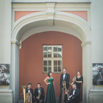 Zespół Masterbrass, ubrany elegancko z zielonymi dodatkami, trzymający swoje instrumenty, z sopranistką ubraną w długą zielono-czarną suknię, usytuowani na tle okna i dwóch obrazów baletnic, na dziedzińcu Ogólnokształcącej Szkoły Baletowej im. Olgi Sławskiej Lipczyńskiej.