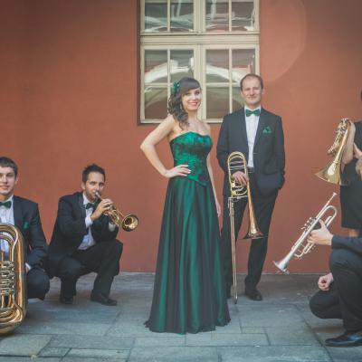 Zespół Masterbrass, ubrany elegancko z zielonymi dodatkami, trzymający swoje instrumenty, jeden trębacz udaje, że gra na trąbce. Jest z nimi sopranistka ubrana w długą zielono-czarną suknię, usytuowani na tle okna dziedzińca Ogólnokształcącej Szkoły Baletowej im. Olgi Sławskiej Lipczyńskiej.