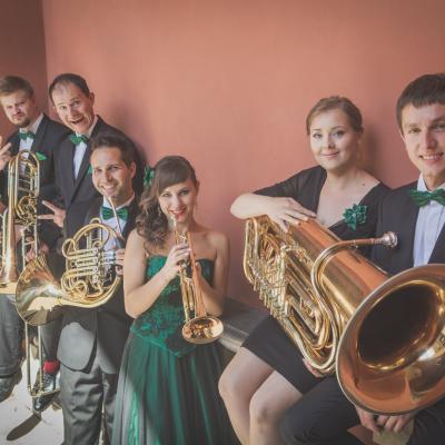Członkowie zespołu Masterbrass, ubrani elegancko z zielonymi dodatkami wraz z solistką ubraną w długą, zielono-czarną suknię, z powymienianymi między sobą instrumentami, śpiewaczka udaje, że gra na trąbce, wszyscy uśmiechnięci w żartobliwym nastroju, stoją a nie którzy siedzą na poręczy schodów, na dziedzińcu Ogólnokształcącej Szkoły Baletowej im. Olgi Sławskiej Lipczyńskiej.