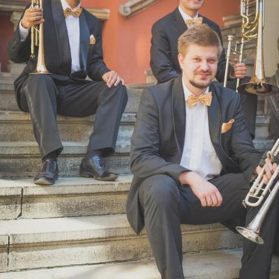 Dwóch trębaczy i puzonista zespołu Masterbrass, ubrani elegancko ze złotymi muszkami, trzymający swoje instrumenty siedząc na schodach dziedzińca Ogólnokształcącej Szkoły Baletowej im. Olgi Sławskiej Lipczyńskiej.