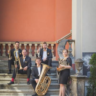 Zespół Masterbrass, ubrany elegancko ze złotymi dodatkami, trzymający swoje instrumenty siedząc, a waltornistka stojąc na schodach dziedzińca Ogólnokształcącej Szkoły Baletowej im. Olgi Sławskiej Lipczyńskiej.