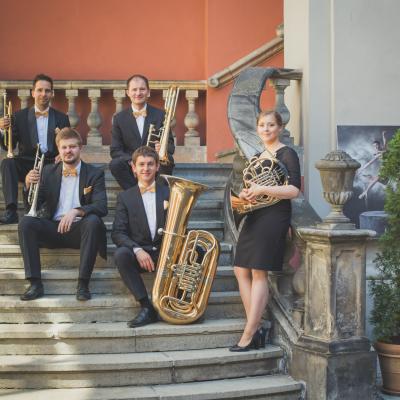Zespół Masterbrass, ubrany elegancko ze złotymi dodatkami, trzymający swoje instrumenty siedząc, a waltornistka stojąc na schodach dziedzińca Ogólnokształcącej Szkoły Baletowej im. Olgi Sławskiej Lipczyńskiej, gdzie po prawej stronie jest obraz baletnicy i zielona tuja.