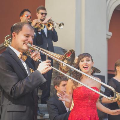 Na pierwszym planie grający puzonista, ubrany w garnitur, ze złotą muszką, a tuż za nim śpiewająca sopranistka, ubrana w czerwoną suknię ze złotym kwiatkiem we włosach. W tle grający zespół Masterbrass, elegancko ubrany ze złotymi dodatkami, trębacze w okularach. Wszyscy stoją na schodach dziedzińca Ogólnokształcącej Szkoły Baletowej im. Olgi Sławskiej Lipczyńskiej.