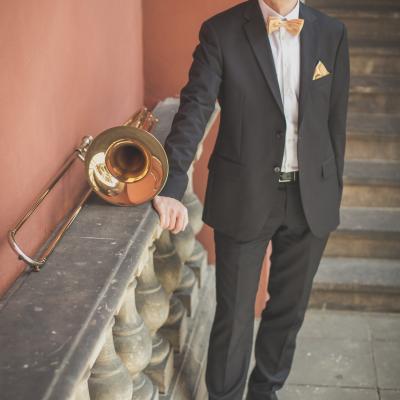 Puzonista, elegancko ubrany ze złotą muszką, stojący ze swoim instrumentem na tle schodów dziedzińca Ogólnokształcącej Szkoły Baletowej im. Olgi Sławskiej Lipczyńskiej.