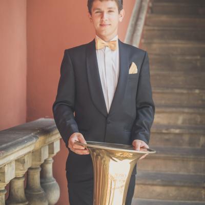 Tubista, elegancko ubrany ze złotą muszką, stojący ze swoim instrumentem na tle schodów dziedzińca Ogólnokształcącej Szkoły Baletowej im. Olgi Sławskiej Lipczyńskiej.