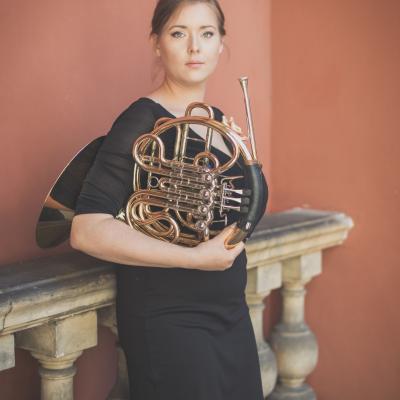 Waltornistka, ubrana w czarną sukięnkę, stojąca ze swoim instrumentem, na tle balustrady dziedzińca Ogólnokształcącej Szkoły Baletowej im. Olgi Sławskiej Lipczyńskiej.