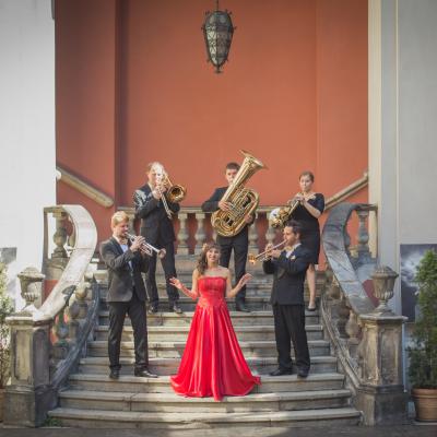 Członkowie zespołu Masterbrass, ubrani elegancko ze złotymi dodatkami wraz z solistką ubraną w długą, czerwoną suknię, ze złotym kwiatkiem wpiętym we włosy, grający na schodach dziedzińca Ogólnokształcącej Szkoły Baletowej im. Olgi Sławskiej Lipczyńskiej.