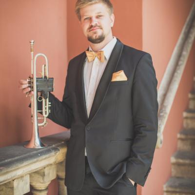 Trębacz, elegancko ubrany ze złotą muszką, stojący przy balustradzie, opierając na niej swój instrument, na dziedzińcu Ogólnokształcącej Szkoły Baletowej im. Olgi Sławskiej Lipczyńskiej.