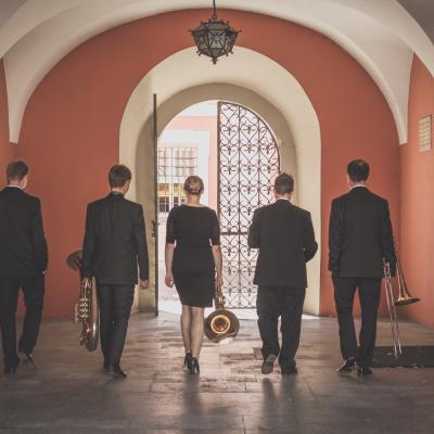 Zespół Masterbrass, ubrany elegancko, idący ze swoimi instrumentami w stronę czarnej bramy wyjściowej z Ogólnokształcącej Szkoły Baletowej im. Olgi Sławskiej Lipczyńskiej.