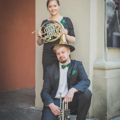 Waltornistka trzymająca waltornię obróconą czarą na głowie przykucniętego trębacza, na dziedzińcu Ogólnokształcącej Szkoły Baletowej im. Olgi Sławskiej Lipczyńskiej.