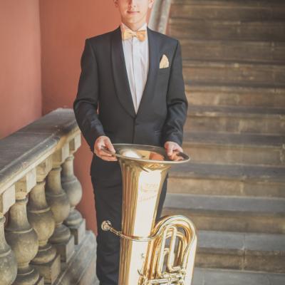 Tubista zespołu Masterbrass, elegancko ubrany w garnitur ze złotą muszką wraz ze swoją Tubą, stoi na tle schodów, na dziedzińcu Ogólnokształcącej Szkoły Baletowej im. Olgi Sławskiej Lipczyńskiej.