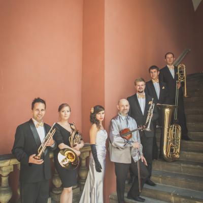 Członkowie zespołu Masterbrass, ubrani elegancko ze złotymi dodatkami ze swoimi instrumentami wraz z solistką ubraną w długą, srebrną suknię, ze złotym kwiatkiem wpiętym we włosy oraz ze skrzypkiem ubranym w srebrną koszulę i czarne spodnie trzymającym swoje skrzypce, stoją na schodach, na dziedzińcu Ogólnokształcącej Szkoły Baletowej im. Olgi Sławskiej Lipczyńskiej.