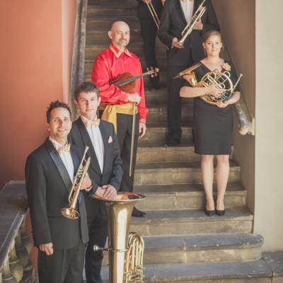 Zespół Masterbrass, ubrany elegancko ze złotymi muszkami, trzymający swoje instrumenty, i skrzypek w czerwonej koszuli i czarnych spodniach, trzymający swój instrument, stoją na schodach dziedzińca Ogólnokształcącej Szkoły Baletowej im. Olgi Sławskiej Lipczyńskiej.