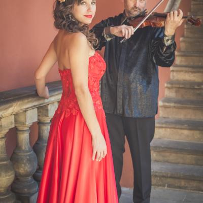 Sopranistka w czerwonej, długiej sukni z wpiętym złotym kwiatkiem we włosy, patrząca w dal, a w tle grający skrzypek, ubrany na czarno, spoglądający na nią, stoją na dziedzińcu Ogólnokształcącej Szkoły Baletowej im. Olgi Sławskiej Lipczyńskiej.