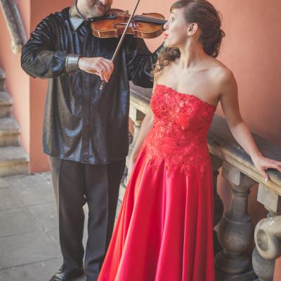 Sopranistka w czerwonej, długiej sukni i grający skrzypek, ubrany na czarno, muzykują w patrzeni sobie w oczy, na dziedzińcu Ogólnokształcącej Szkoły Baletowej im. Olgi Sławskiej Lipczyńskiej.