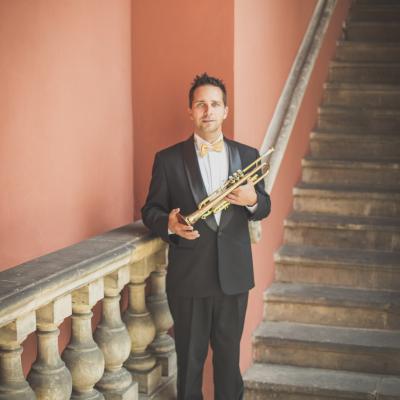 Trębacz, elegancko ubrany ze złotą muszką, stojący ze swoim instrumentem na tle schodów dziedzińca Ogólnokształcącej Szkoły Baletowej im. Olgi Sławskiej Lipczyńskiej.