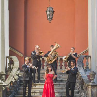 Członkowie zespołu Masterbrass, ubrani elegancko ze złotymi dodatkami wraz z solistką ubraną w długą, czerwoną suknię, ze złotym kwiatkiem wpiętym we włosy, grający na schodach dziedzińca Ogólnokształcącej Szkoły Baletowej im. Olgi Sławskiej Lipczyńskiej.