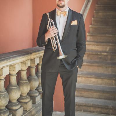 Trębacz, elegancko ubrany ze złotą muszką, stojący ze swoim instrumentem na tle schodów dziedzińca Ogólnokształcącej Szkoły Baletowej im. Olgi Sławskiej Lipczyńskiej.