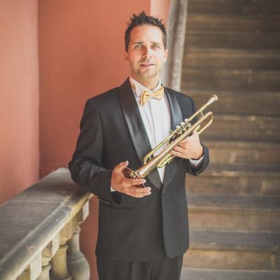 Trębacz, elegancko ubrany ze złotą muszką, stojący ze swoim instrumentem na tle schodów dziedzińca Ogólnokształcącej Szkoły Baletowej im. Olgi Sławskiej Lipczyńskiej.