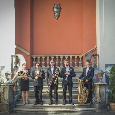 Zespół Masterbrass, ubrany elegancko ze złotymi dodatkami, trzymający swoje instrumenty stojąc na jednym stopniu schodów dziedzińca Ogólnokształcącej Szkoły Baletowej im. Olgi Sławskiej Lipczyńskiej, gdzie po prawej i lewej stronie jest obraz baletnicy i zielona tuja.