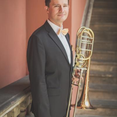 Puzonista, elegancko ubrany ze złotą muszką, stojący bokiem ze swoim instrumentem na tle schodów dziedzińca Ogólnokształcącej Szkoły Baletowej im. Olgi Sławskiej Lipczyńskiej.