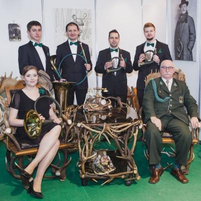 Zespół Masterbrass w większości stojący, waltornistka siedząca na pięknym krześle z porożami, wszyscy z rogami myśliwskimi w pokoju myśliwskim wraz z Krzysztofem Kadlecem