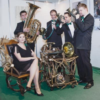 Członkowie zespołu Masterbrass grający na rogach myśliwskich i Tubie, dla waltornistki, siedzącej na ozdobnym krześle z porożami w pokoju myśliwskim.