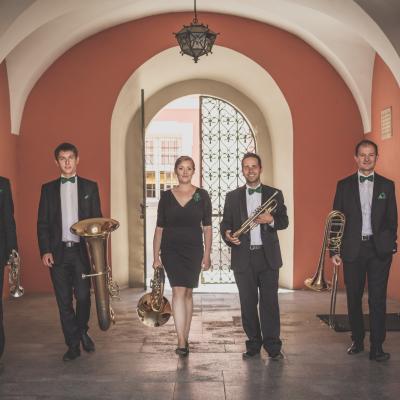 Zespół Masterbrass, ubrany elegancko z zielonymi dodatkami, idący do przodu ze swoimi instrumentami, na tle czarnej bramy wejściowej do Ogólnokształcącej Szkoły Baletowej im. Olgi Sławskiej Lipczyńskiej.