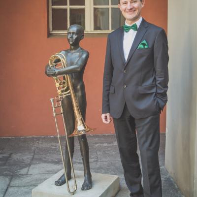 Posąg baletmistrza trzymający ręce w pierwszej pozycji z puzonem a obok niego stoi Nasz puzonista w eleganckim garniturze z zieloną muszką na dziedzińcu Ogólnokształcącej Szkoły Baletowej im. Olgi Sławskiej Lipczyńskiej.