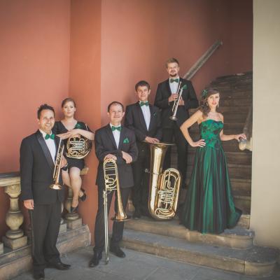 Członkowie zespołu Masterbrass, ubrani elegancko z zielonymi dodatkami ze swoimi instrumentami, wraz z solistką ubraną w długą, zielono-czarną suknię stoją na schodach, na dziedzińcu Ogólnokształcącej Szkoły Baletowej im. Olgi Sławskiej Lipczyńskiej.