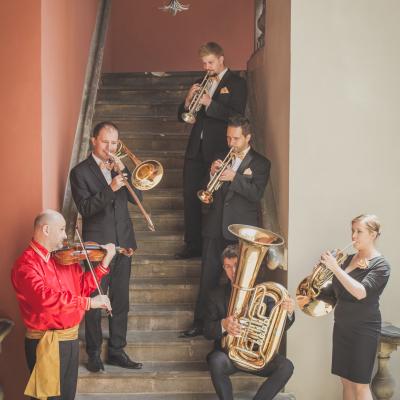 Zespół Masterbrass, ubrany elegancko ze złotymi muszkami oraz skrzypek w czerwonej koszuli i czarnych spodniach, grają na instrumentach, na schodach dziedzińca Ogólnokształcącej Szkoły Baletowej im. Olgi Sławskiej Lipczyńskiej.
