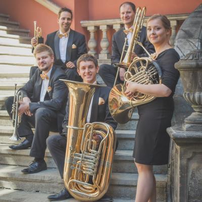 Zespół Masterbrass, ubrany elegancko ze złotymi dodatkami, trzymający swoje instrumenty siedząc na schodach dziedzińca Ogólnokształcącej Szkoły Baletowej im. Olgi Sławskiej Lipczyńskiej.